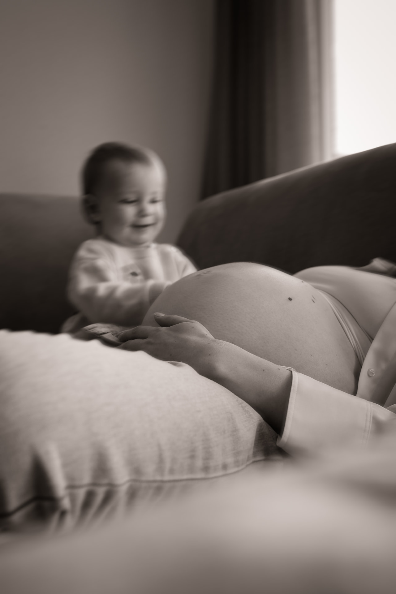 Schwarzweiß-Aufnahme: Kleinkind sitzt lachend neben dem Babybauch der Mutter auf dem Sofa