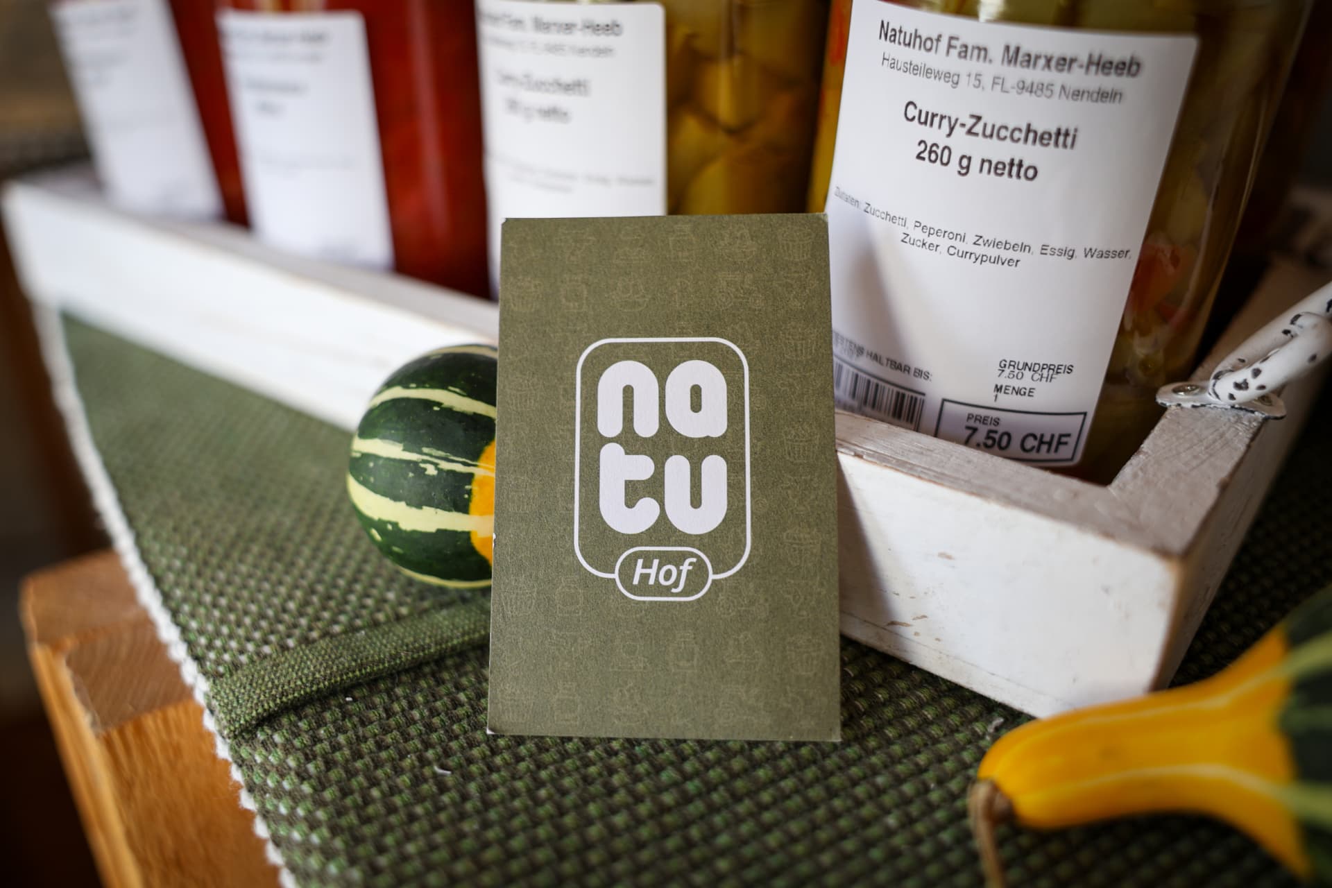 Olivgrüne Natu-Hof-Karte steht im Hofladen vor Gläsern mit Curry-Zucchetti